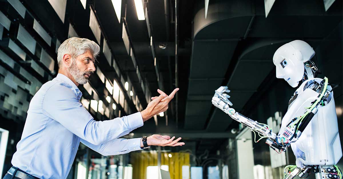 A man gestures with his hands while facing a humanoid robot—someone using AI—in a modern, industrial-style building.