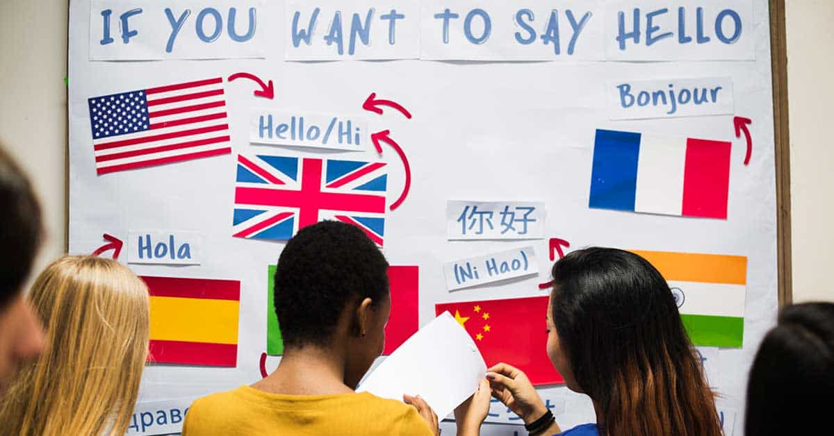 A group of people stand in front of a poster showing how to say "hello" in multiple languages, including English, Spanish, Chinese, French, and others—an empowering display promoting bilingualism for new immigrant students.