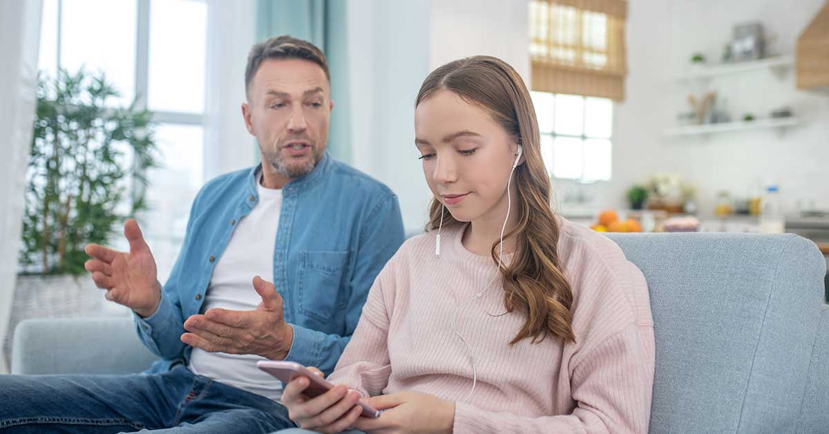 A man talks to a girl who is listening to something with earphones and looking at her phone, while sitting together on a couch in a living room, as they discuss the dangers of sextortion online.