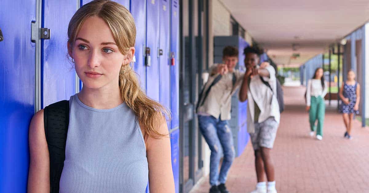 A teenage girl stands alone by lockers looking sad while two boys in the background point and laugh at her in a school hallway, hinting at the pain caused by sextortion and online bullying.