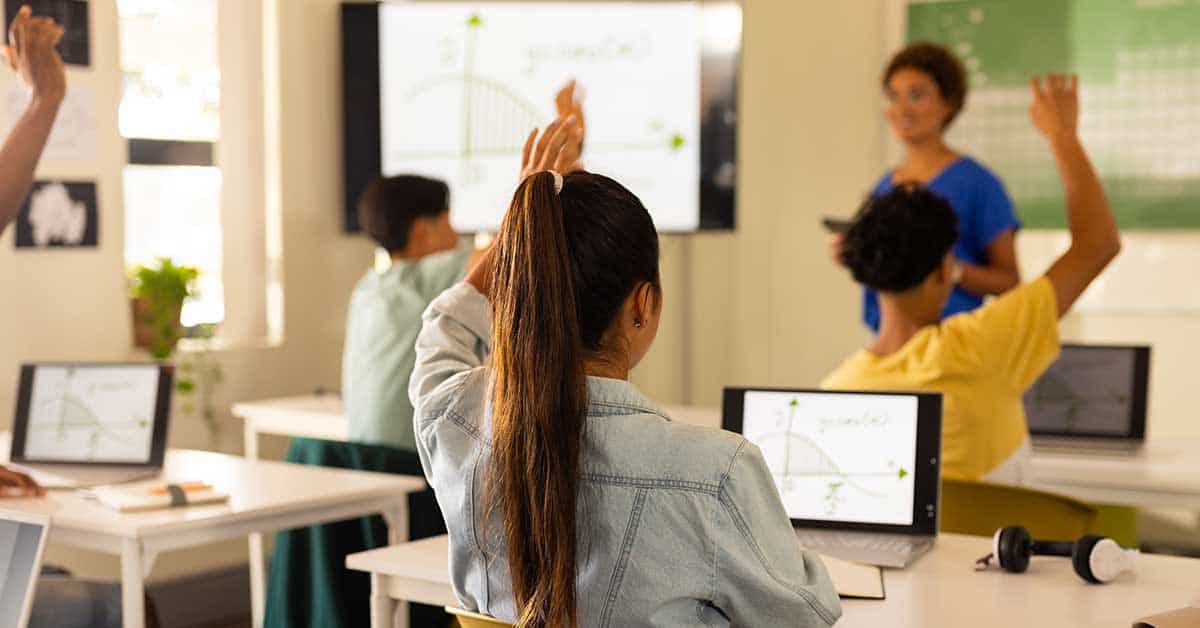 In a dynamic classroom, students eagerly raise their hands to answer questions, while laptops flashing graphs sit on desks. At the front, the teacher guides exploration with AI-powered learning tools displayed on a screen showing mathematical content.