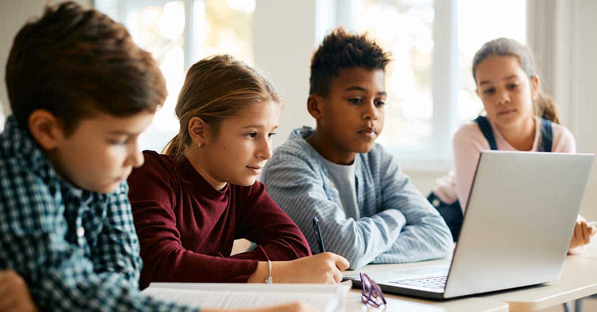 Four students sit at a table, looking intently at a laptop screen and taking notes, with notebooks and pens in front of them.