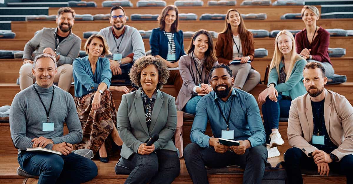 A group of twelve professionally dressed adults with conference badges sit on wooden steps in an auditorium, smiling at the camera, representing a dynamic system of collaboration and expertise.