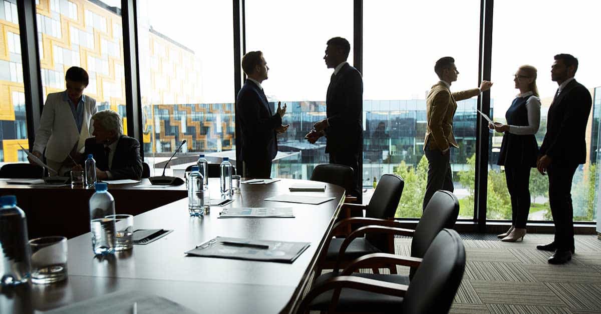 People standing and talking in a modern conference room with a long table, chairs, and water bottles, exuding a sense of leadership, next to large windows overlooking an office building.