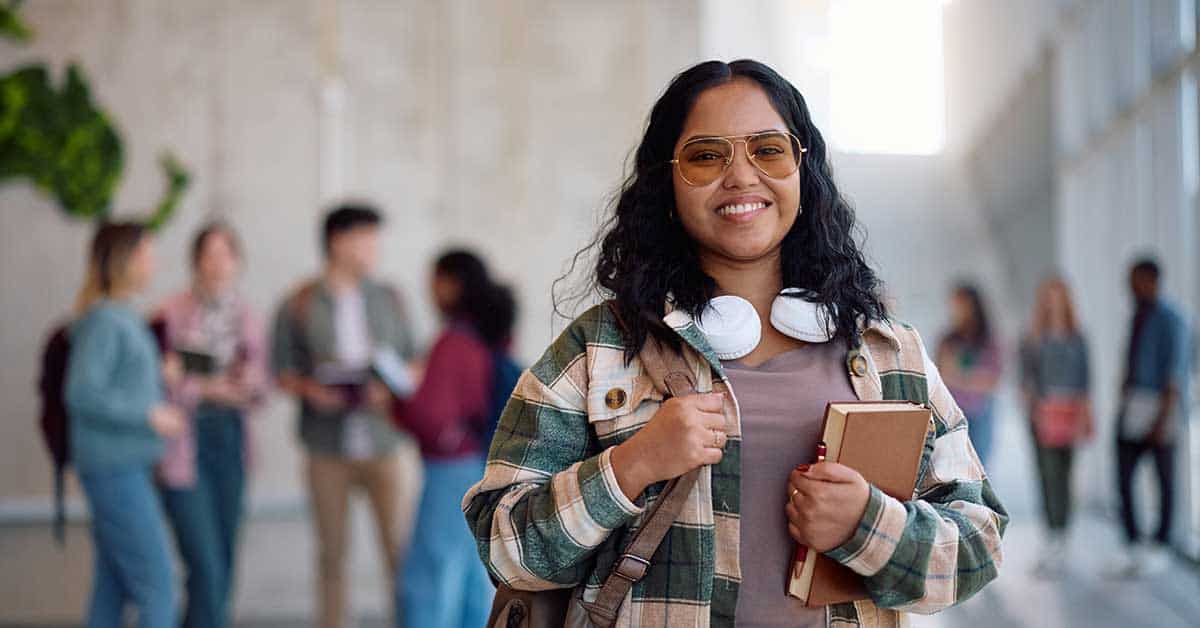 A student wearing glasses and headphones labeled "BIE" around her neck holds books and smiles in a hallway, with a group of people talking in the background.
