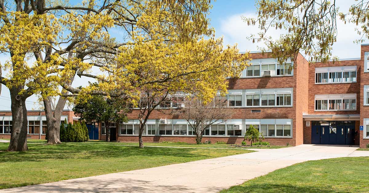 A brick school building with large windows sits amid green grass and trees with yellow leaves, under a partly cloudy sky—an inviting scene for students purchasing books or supplies each morning.