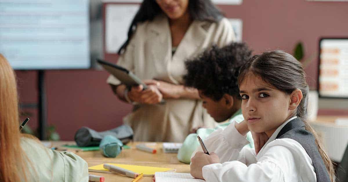 In a classroom bustling with high expectations, a girl looks directly at the camera, holding a pencil and notebook. Behind her, fellow students and a teacher with a tablet are engaged in learning, representing the hopeful future of education reform.