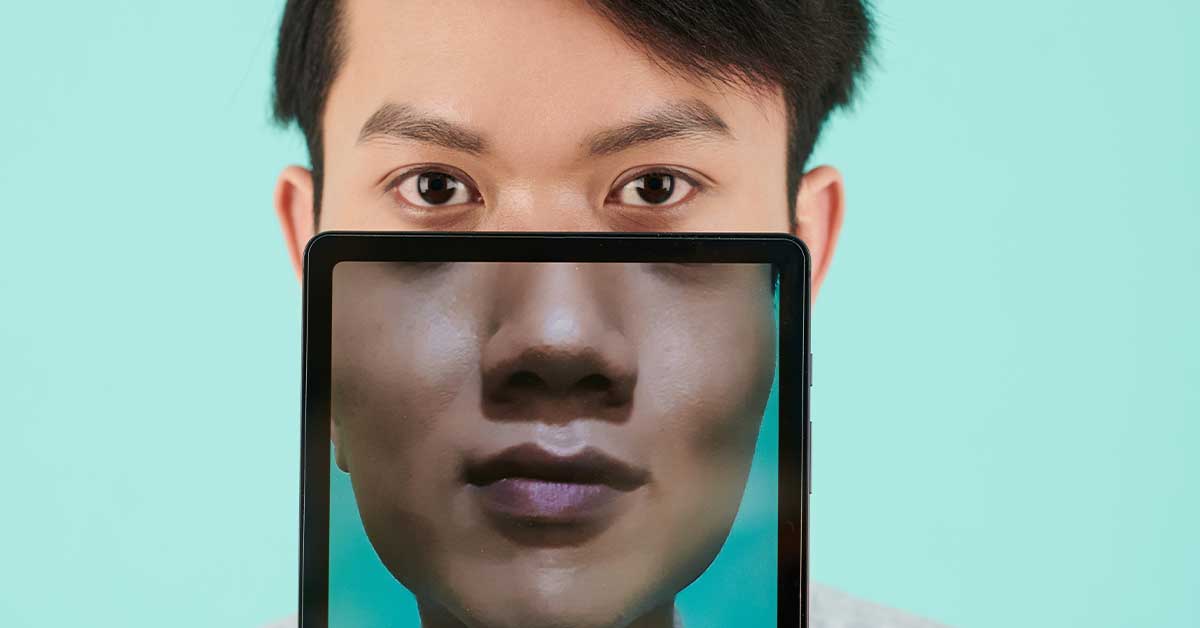 A person holds a tablet in front of their face, displaying a close-up of their lower face—suggesting the potential for deepfake manipulation—while their eyes are visible above the screen against a light blue background.