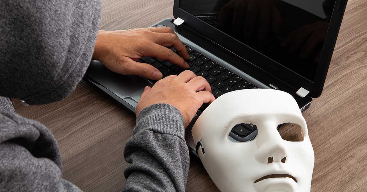 A person in a hoodie types on a laptop at a wooden table, with a white mask—hinting at deepfake deception—resting beside the keyboard.