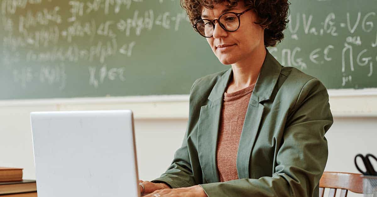 A person wearing glasses and a green blazer types on a laptop in front of a chalkboard filled with mathematical equations, highlighting concerns school leaders face about deepfake blackmail and sextortion threats in educational settings.
