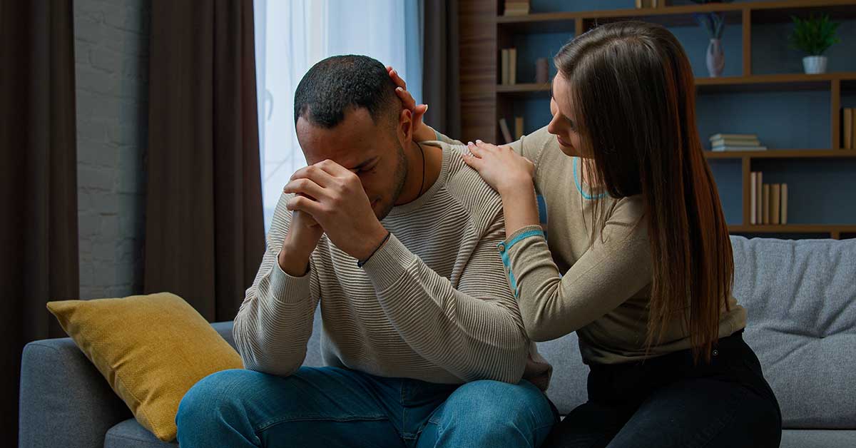A woman sits on a couch comforting a man who appears distressed, possibly dealing with the emotional toll of sextortion, his elbows on his knees and hands clasped near his face.