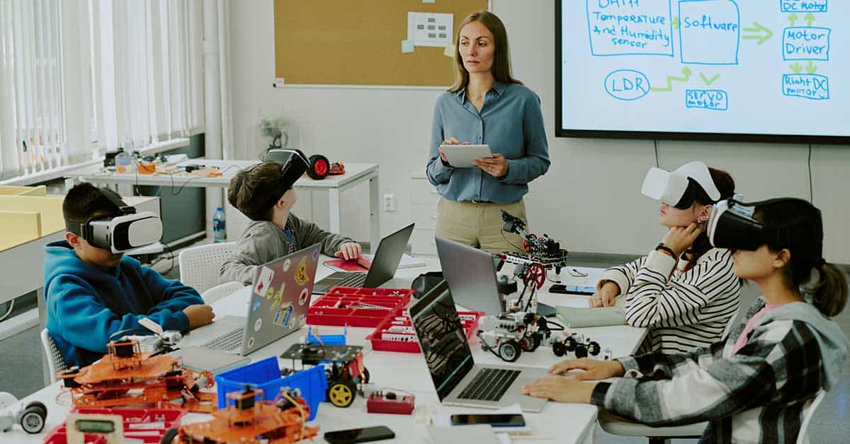 A teacher stands in front of a screen, discussing AI in education with students wearing VR headsets. The students sit at a table, immersed in the lesson, surrounded by laptops and robotics kits in a modern classroom setting.