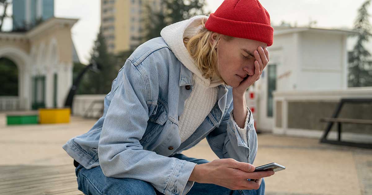 A young person in a red beanie and denim jacket sits outdoors, looking at a smartphone and holding their head in apparent frustration, possibly experiencing cyberbullying.