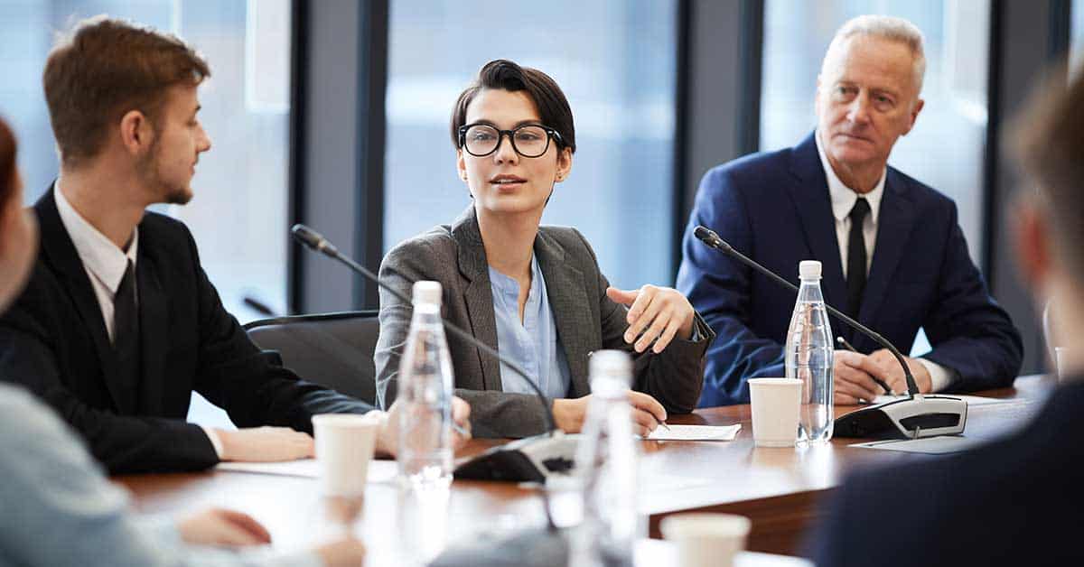 In a business meeting with four people seated at a conference table, a person in glasses addresses the group while others listen intently. Bottles of water and microphones are on the table, setting the stage for discussions that could touch upon hostile boards or dysfunctional governance issues.