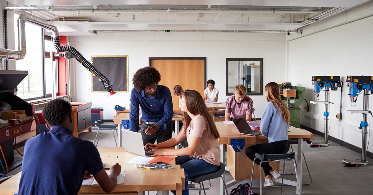A group of students work on laptops and papers in a classroom with machinery, engaging in hands-on education while a teacher assists one student.