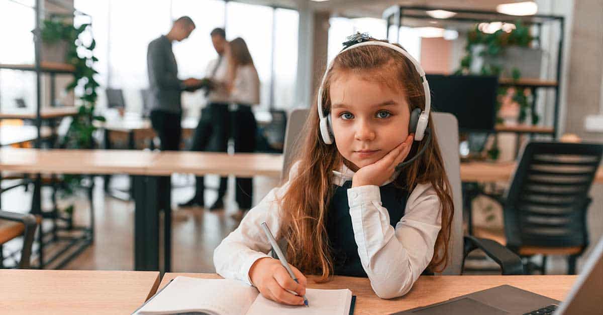 A young girl wearing headphones sits at a desk with a notebook and pen, embodying modern educational practices. She looks at the camera while blurred adults move in the office's background.