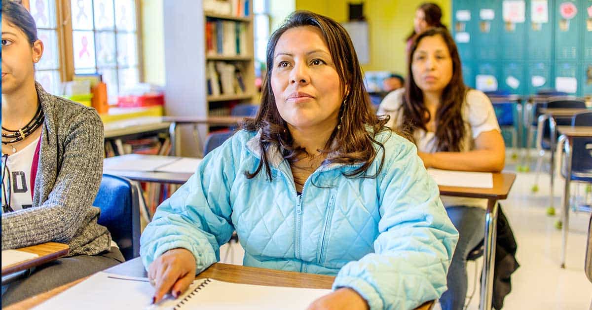 A woman in a blue jacket sits at a desk in a tribal school classroom, attentively looking forward with papers and a pen before her at a tribal school. Other students and elements of this vibrant learning environment are visible in the background.