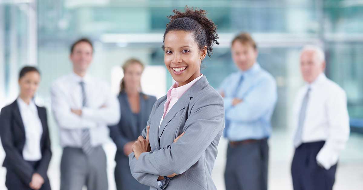A woman in a gray suit stands confidently with her arms crossed, setting high standards and expectations for her professionally dressed colleagues in an office setting.