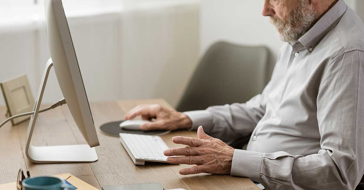 An older man with a gray beard sits at a desk using a desktop computer with a keyboard and mouse, bridging the digital divide.