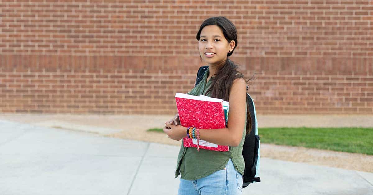 A student with a backpack stands outside in front of a brick wall, holding a red notebook and other BIE school supplies.