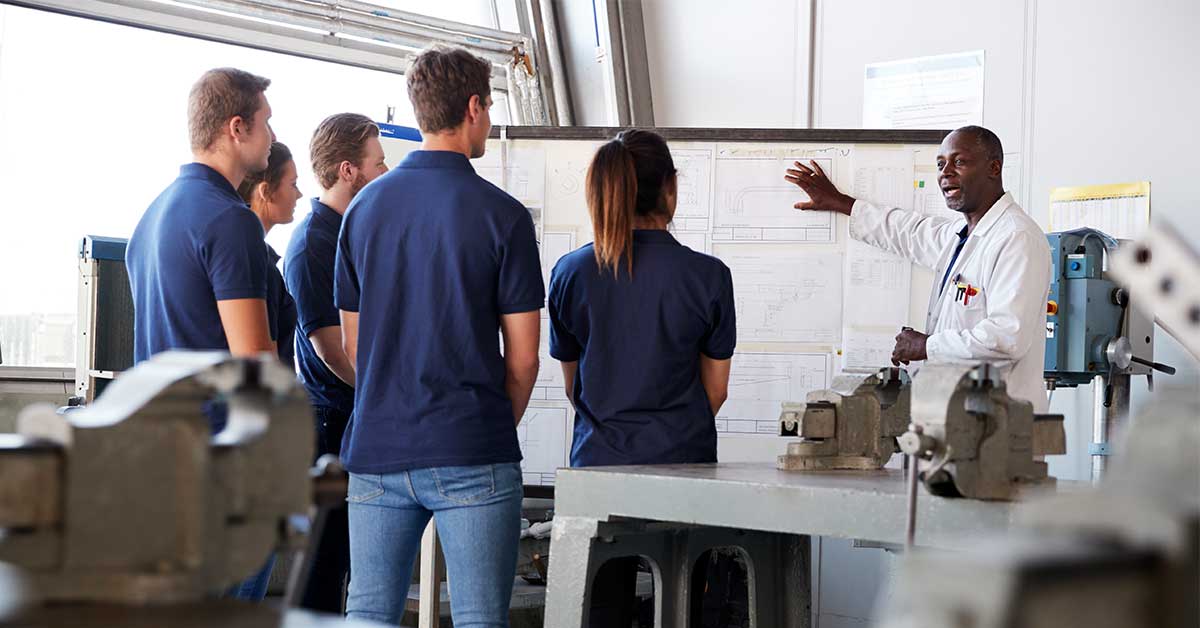 An instructor in a white coat explains technical drawings on a whiteboard to a group of five students wearing blue shirts in a school workshop setting.