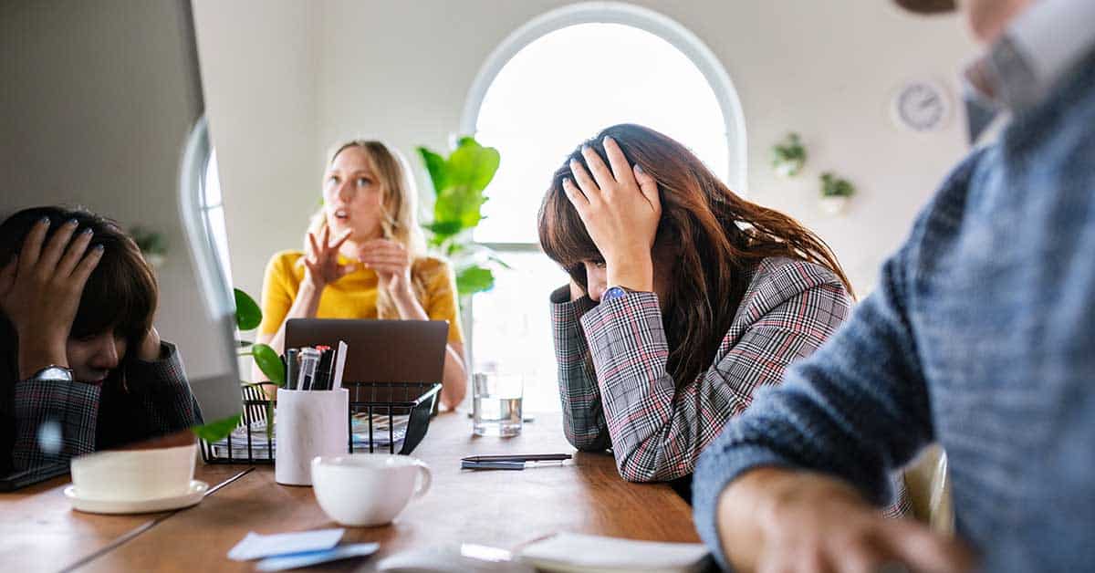 Three people sit at a conference table; one woman, possibly impacted by toxic leadership, has her head in her hands looking stressed, while another woman speaks and a man sits in the foreground.
