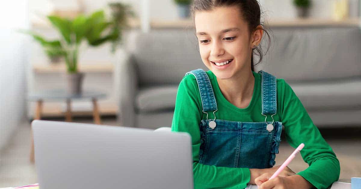 A young girl in a green shirt and denim overalls smiles while homeschooling, using a laptop and writing in a notebook at a table in a living room.