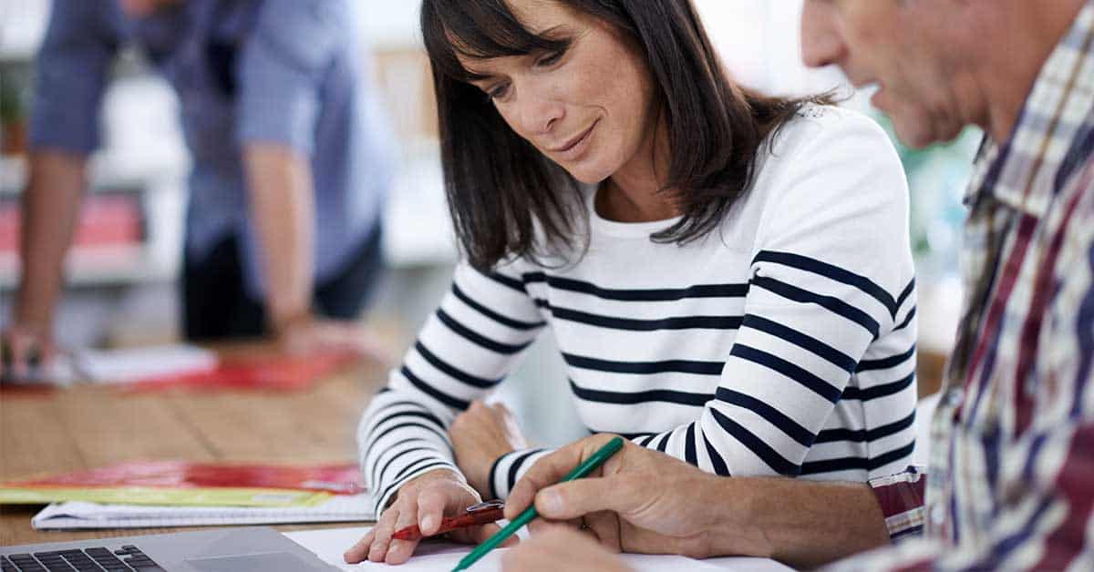 Two people sit at a table collaborating on a grant application, writing on paper with pencils, while a laptop and documents are nearby. A third person stands in the background.
