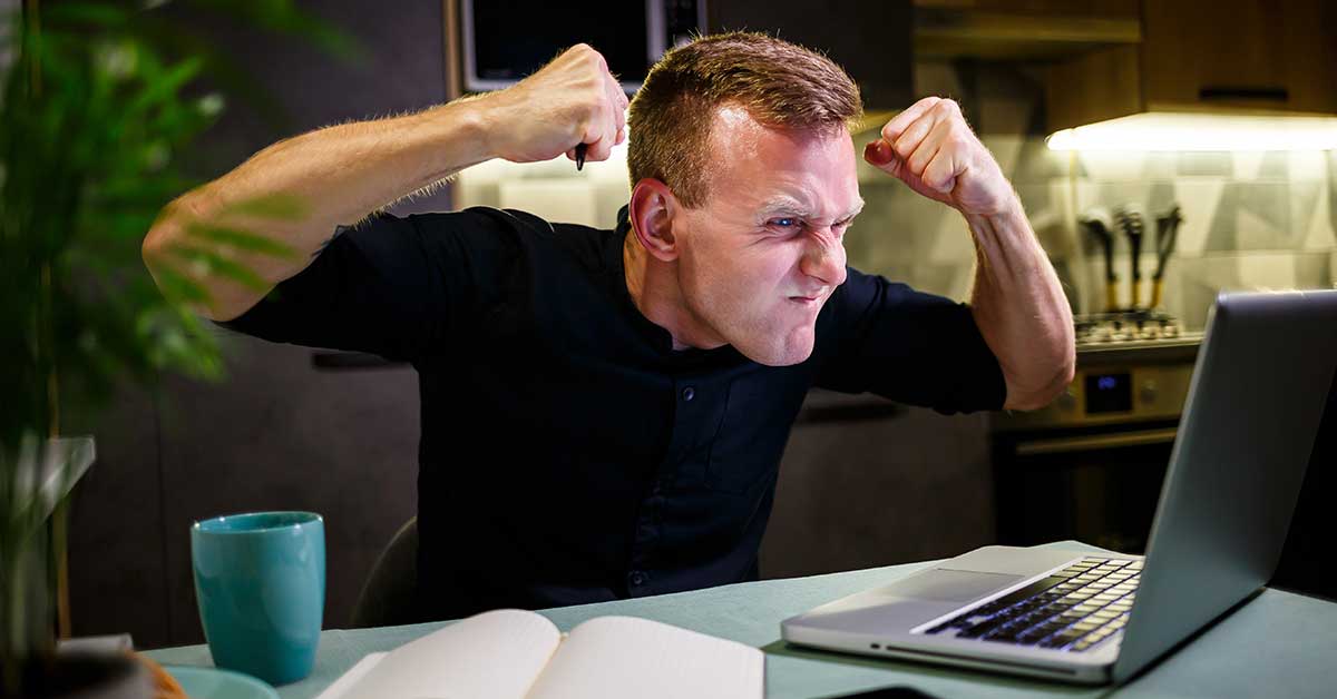 A man sits at a table in front of a laptop, clenching his fists and making an angry facial expression, possibly reacting to content linked to radicalization.