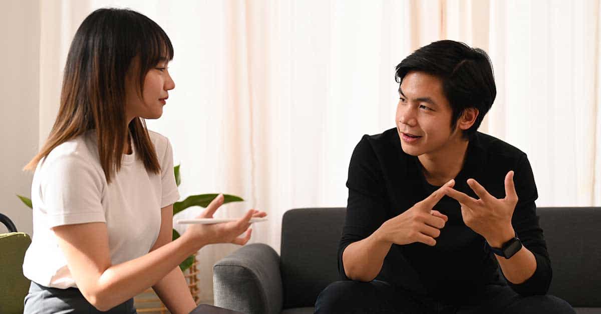 Two people sit on a couch having a conversation about love languages. The woman gestures with her hands while the man counts on his fingers. A plant is visible in the background.