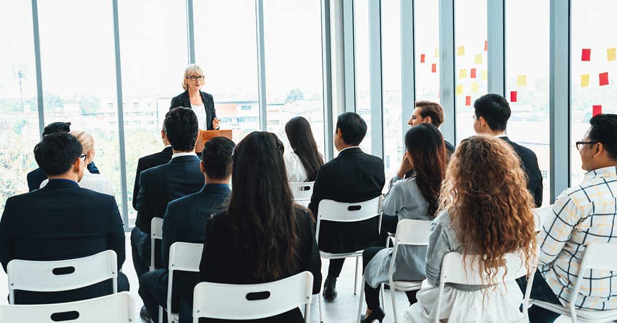 A person stands at a podium addressing a seated audience in a bright room with large windows and sticky notes on the glass, discussing micro-credentials and their significance in our AI-driven world.