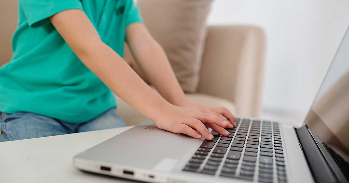 A child wearing a green shirt is using a laptop on a white table in a living room setting, highlighting concerns about online exploitation.