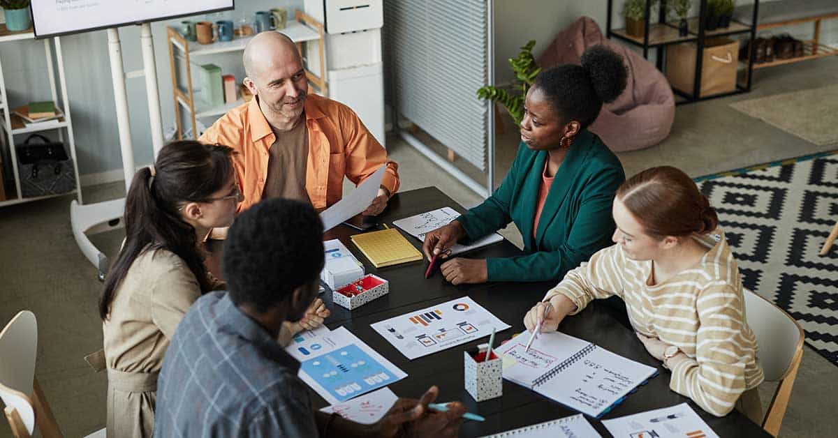 Five people sit around a table in an office, engaging in an organizational meeting as they discuss documents and charts.