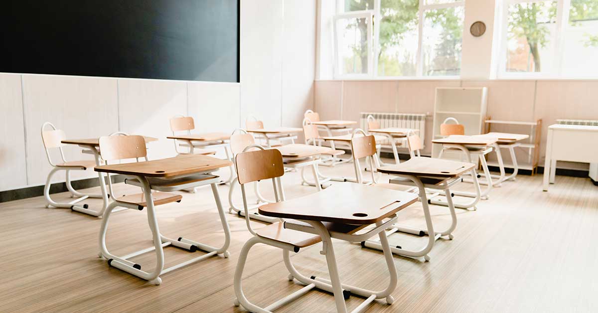 Empty classroom with wooden desks and chairs arranged in rows, large windows filling the BIE learning space with natural light, and a blackboard at the front.