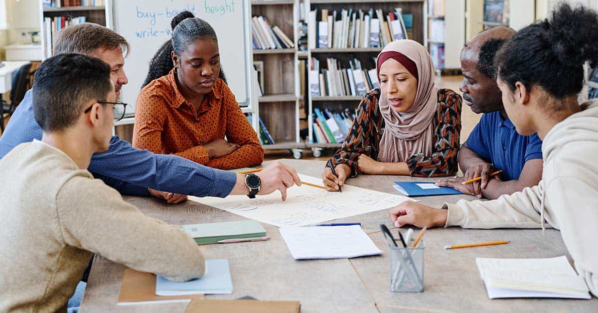 A diverse group of six people sit around a table in a library, discussing and writing on a large sheet of paper. Their focus is addressing academic deficiencies through innovative strategies in early childhood education.