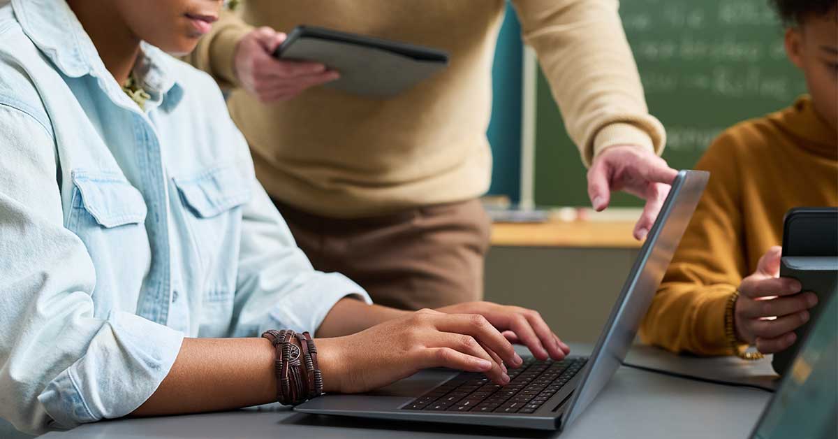 A person types on a laptop while another points at the screen and a third holds a tablet in a classroom setting, discussing online safety topics like sextortion.