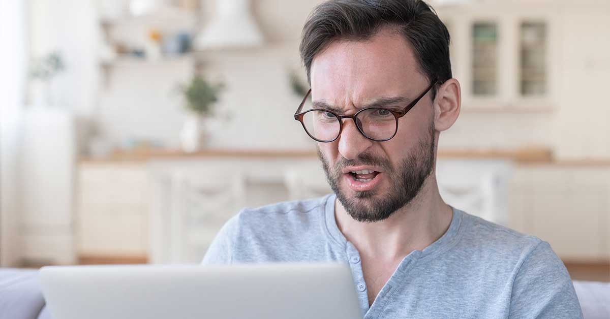 Man with glasses sitting indoors, looking at a laptop with a confused or frustrated expression, his face showing hints of rage as he struggles to understand what’s on the screen.