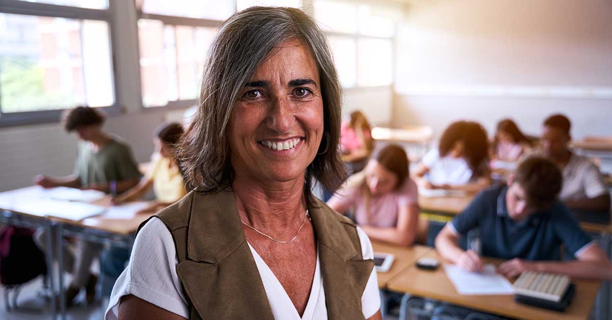 A middle-aged woman stands smiling in a classroom, demonstrating leadership as students sit at desks in the background, working on assignments.
