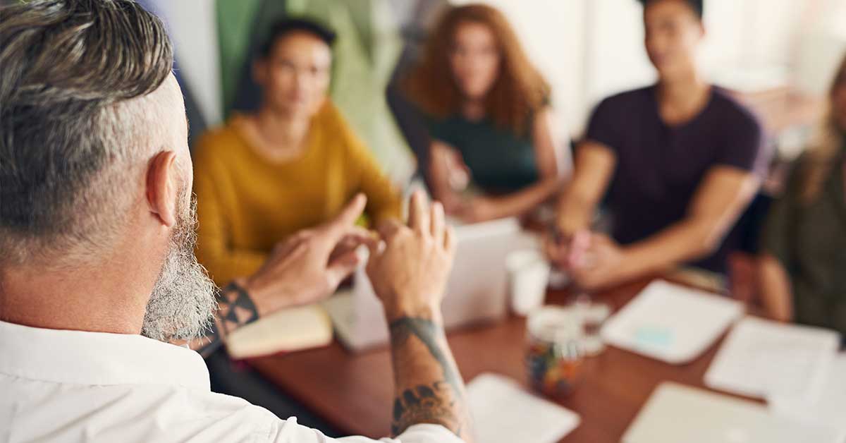 A man with gray hair and a beard, embodying grit, gestures while speaking to a group of four people seated around a table with papers and laptops.