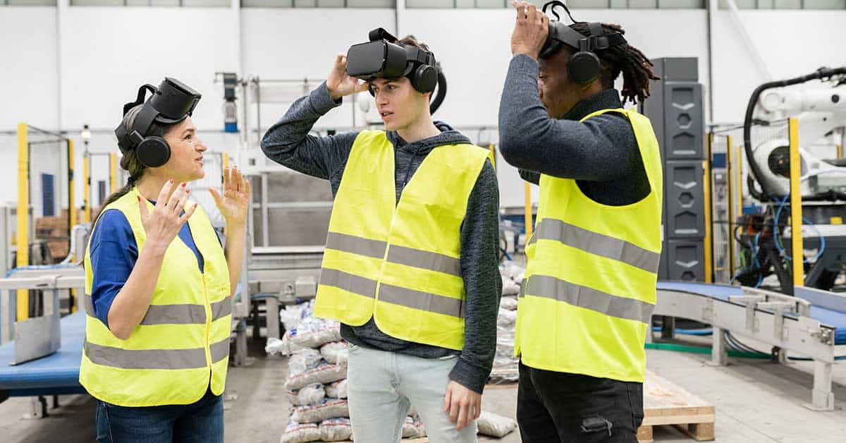 Three people in safety vests and VR headsets stand in a manufacturing facility, appearing to discuss or take off the VR devices.