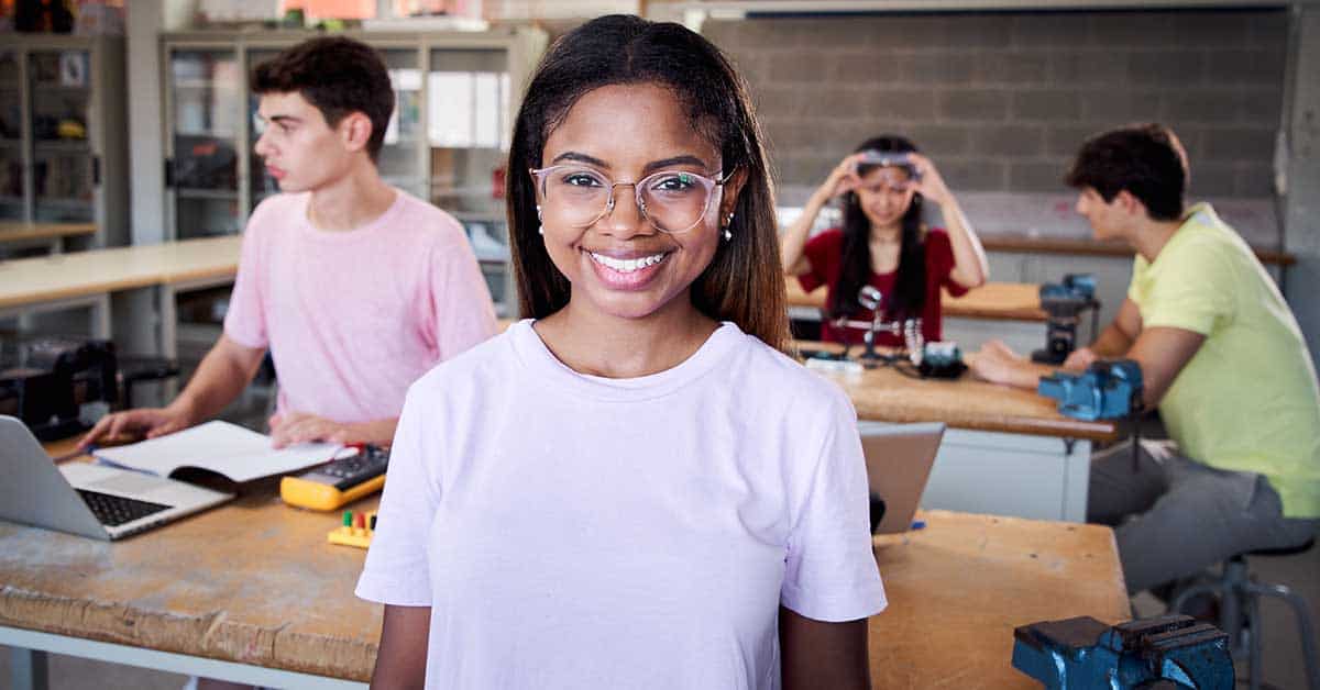 A young woman in glasses stands smiling in the foreground of a classroom, ready to take on the industry, while three other students work on projects at tables in the background.