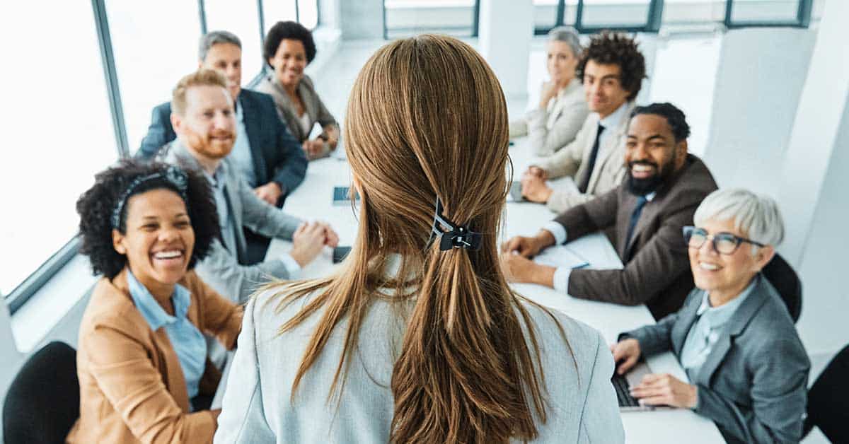 A woman confidently leads a leadership development meeting at the head of a conference table, with a diverse group of nine adults seated around it, all looking attentive.