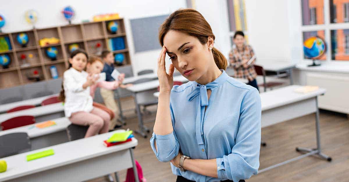 A teacher stands in a rural classroom, looking stressed amidst the chaos of misbehaving children, reflecting the broader staffing crisis faced by many schools today.