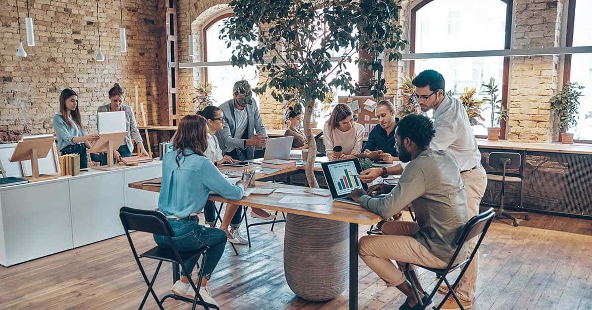 A group of people work together in a modern, open-plan office with large windows, wooden floors, and exposed brick walls. Enjoying a 4-day workweek, some are seated at tables with laptops while others stand and collaborate.