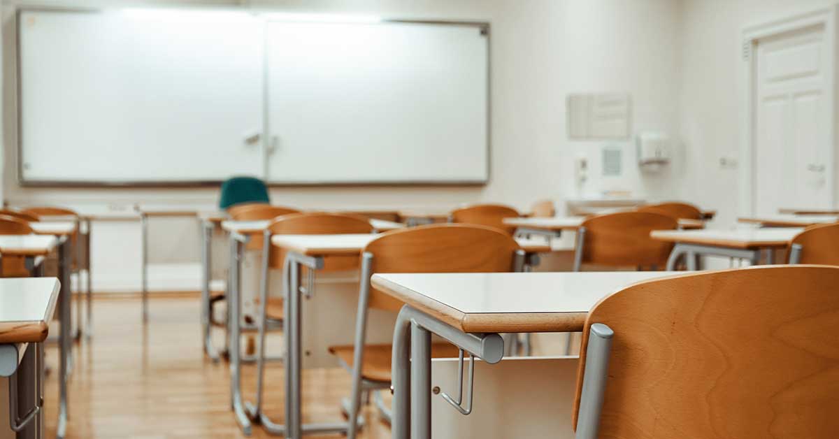 Rows of empty desks and chairs in a well-lit classroom highlight the impact of the teaching shortage, with no instructor present at the whiteboard.