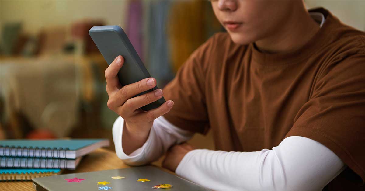 A person in a brown shirt sits at a desk looking at a digital smartphone, with notebooks and a laptop nearby.