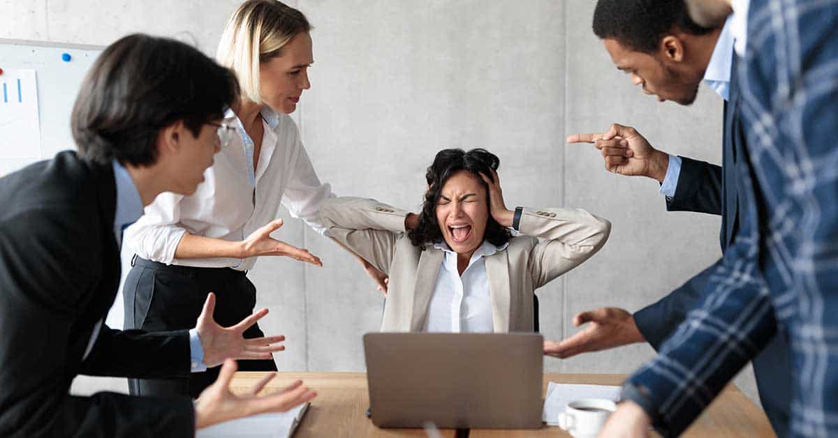 A woman sits at a desk with a laptop, covering her ears in frustration as four colleagues surround her, gesturing with hands—a scene reminiscent of dysfunctional governing boards trying to address issues without reaching a consensus.