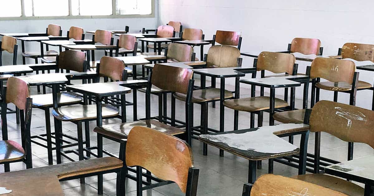 Rows of empty wooden desks and chairs are arranged in a classroom with tiled floors and windows along one wall, awaiting students for their next digital learning session.