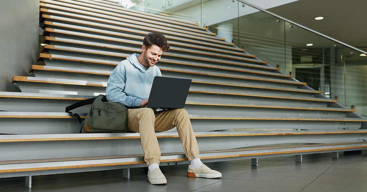 A man sits on indoor stairs, working on a laptop with a bag beside him, in a modern building with glass railings, highlighting the digital divide that access to technology can create in contemporary workspaces.