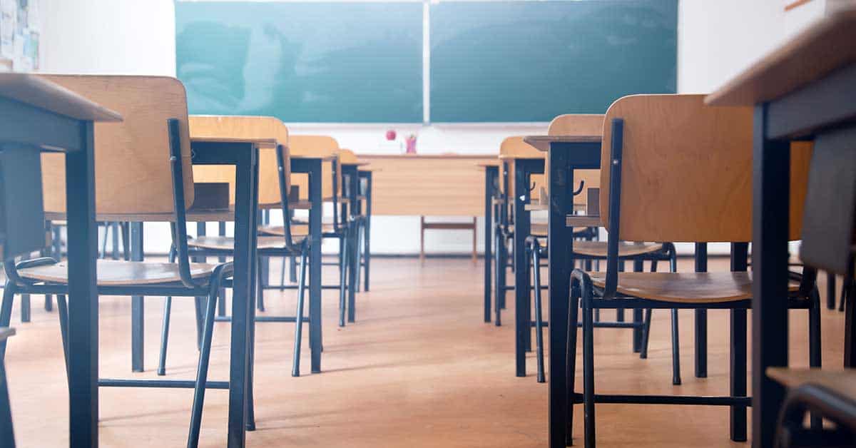 Rows of empty wooden desks and chairs in a classroom, once filled with students from diverse backgrounds shaped by immigration, face a teacher's desk and a green chalkboard at the front of the room.